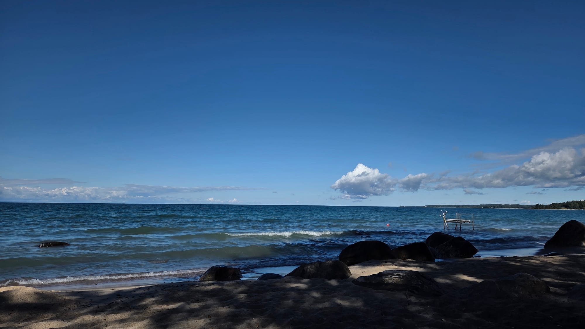 An image of a shady, slightly rocky beach that faces out to the very blue waters of Georgian Bay, the sky is very blue and there's clouds in the distance
